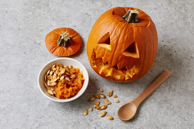 Carved jack-o-lantern pumpkin with triangle eyes and jagged mouth on concrete surface beside bowl of seeds and pulp, wooden spoon nearby