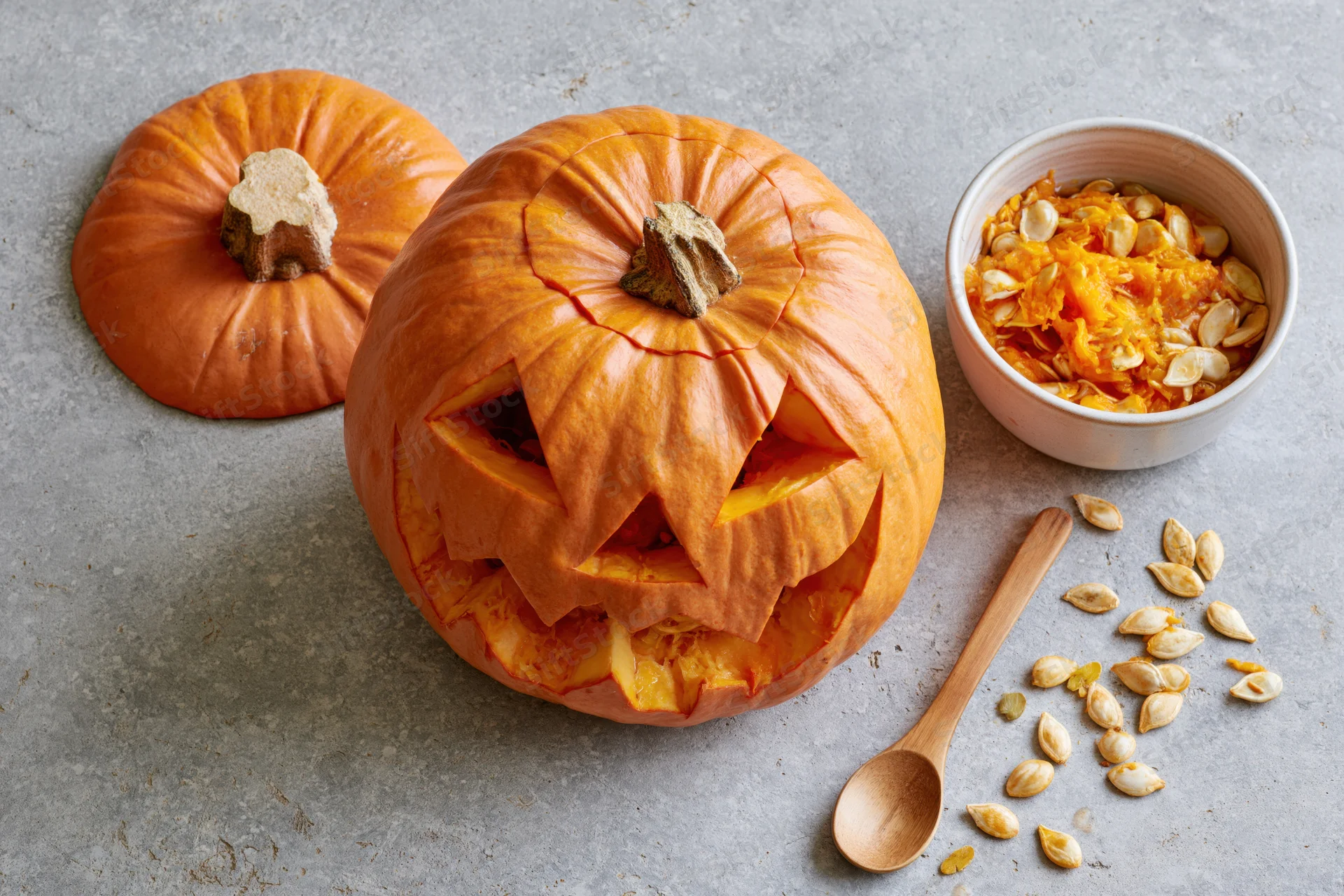 Carved jack-o-lantern pumpkin with triangle eyes and mouth on gray surface beside bowl of seeds and wooden spoon