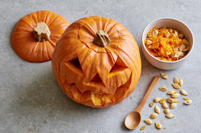 Carved jack-o-lantern pumpkin with triangle eyes and mouth on gray surface beside bowl of seeds and wooden spoon