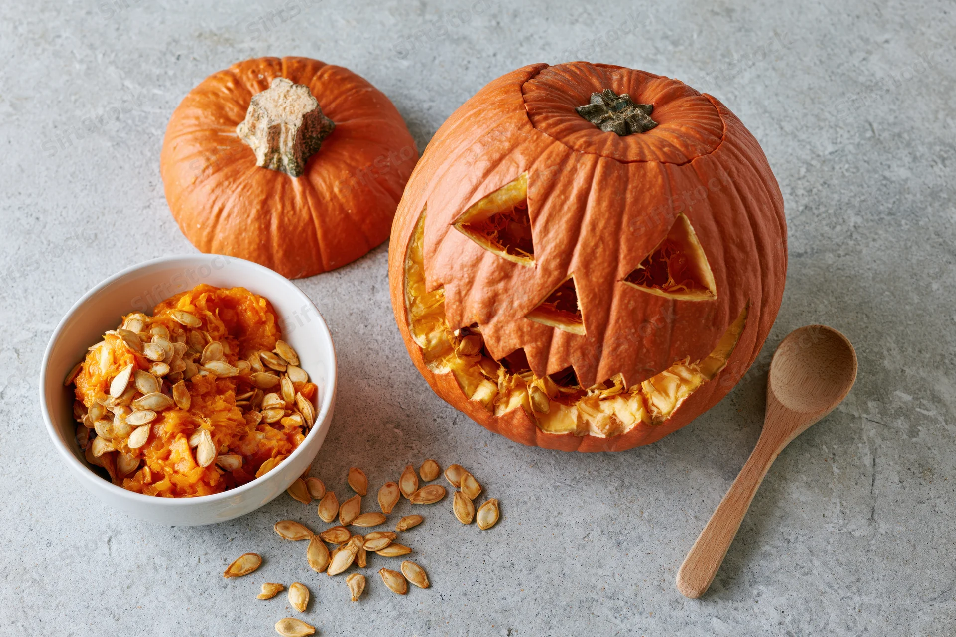 Carved pumpkin with triangle eyes and mouth on gray surface beside bowl of seeds and wooden spoon