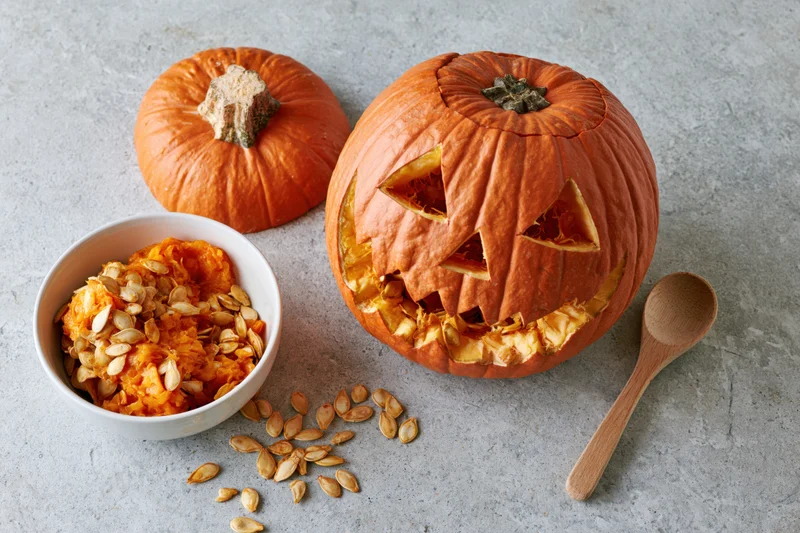 Carved pumpkin with triangle eyes and mouth on gray surface beside bowl of seeds and wooden spoon