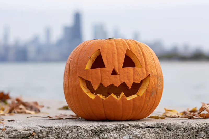 Carved pumpkin with jack-o-lantern face on concrete ledge by lake with city skyline in background