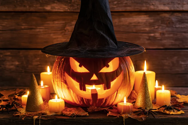 Carved jack-o-lantern with glowing menacing grin wearing witch hat surrounded by candles and autumn leaves on wooden table