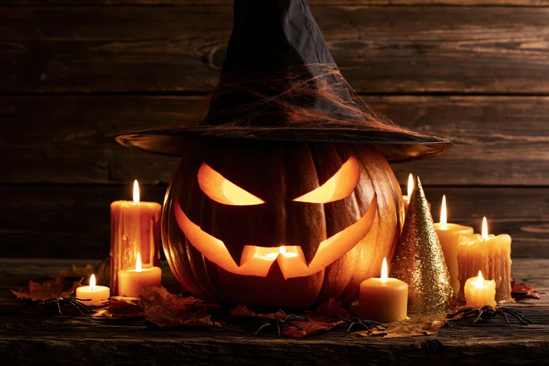 Carved jack-o-lantern with glowing menacing grin wearing witch hat surrounded by lit candles and autumn leaves on wooden table