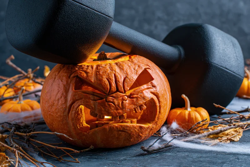 Carved pumpkin with grimacing face under dumbbell on dark surface surrounded by twigs and cobwebs
