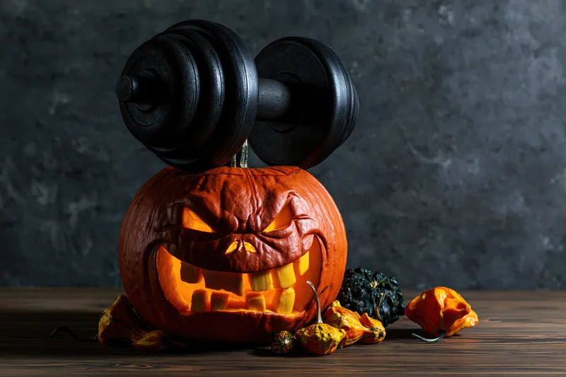 Carved pumpkin with grimacing toothy face and dumbbell on wooden table against dark textured wall