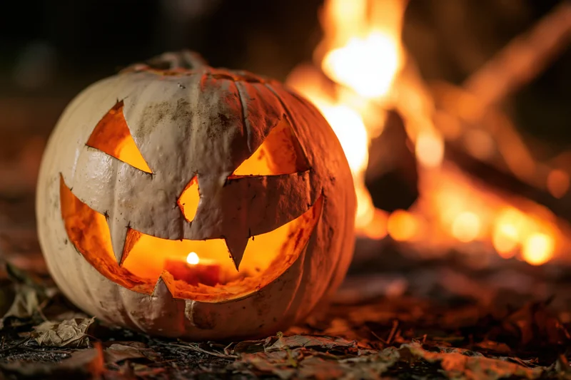 Carved white pumpkin with glowing triangular eyes and jagged grin lit by candle, set on leaf-covered ground near a crackling fire at night
