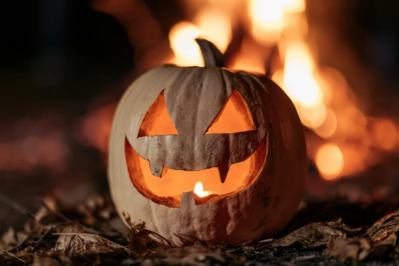 Carved jack-o-lantern glowing with candlelight on dry leaves near fire at night