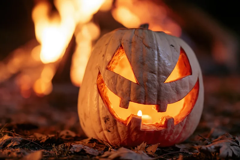 Carved white pumpkin with glowing triangular eyes and wide grin lit by candle, resting on fallen leaves near fire at night