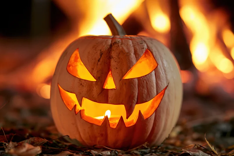 Carved pumpkin with glowing triangular eyes and jagged grin lit by candlelight in front of a fire at night