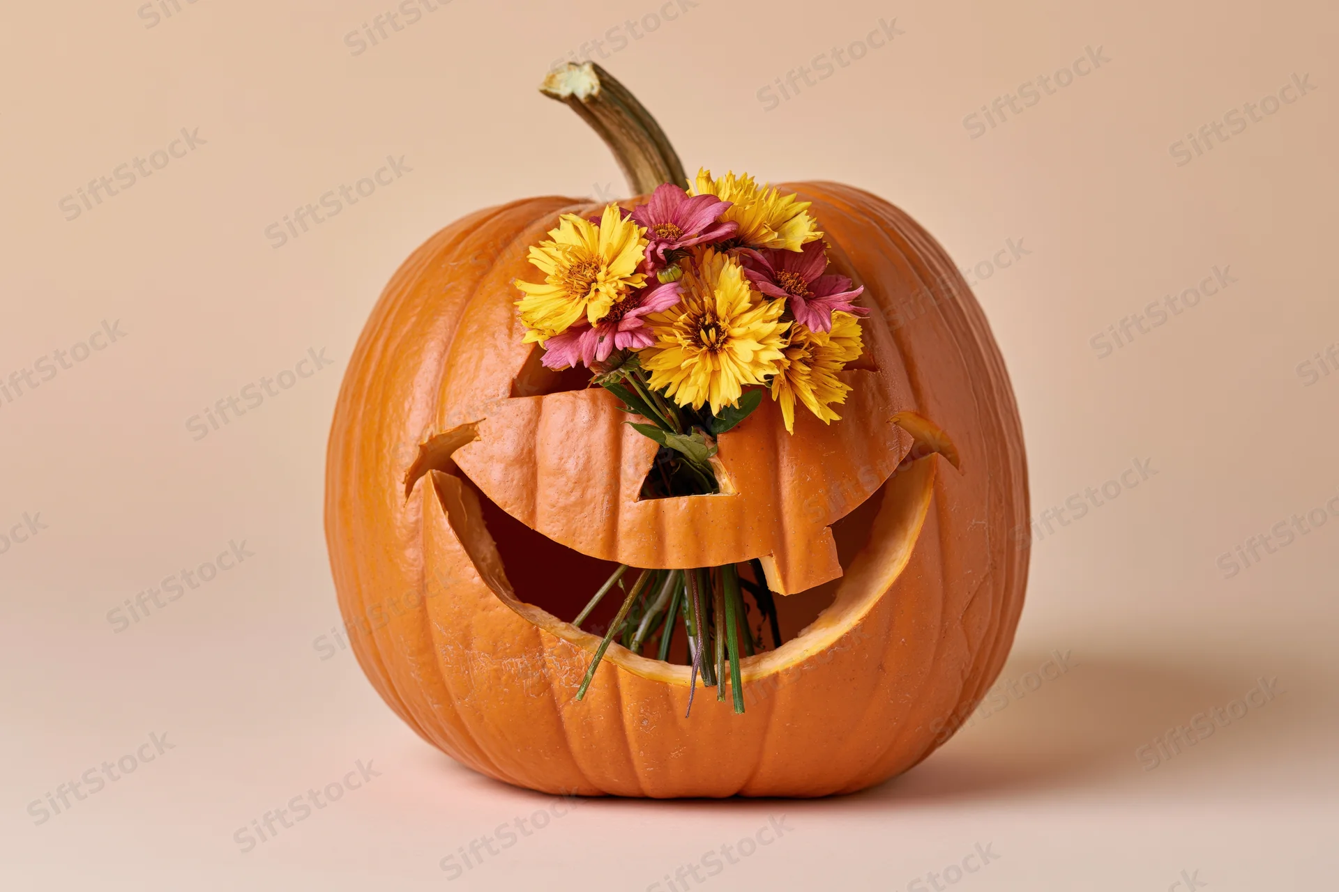 Carved pumpkin with a smiling face holding a bouquet of yellow and pink flowers against a beige background
