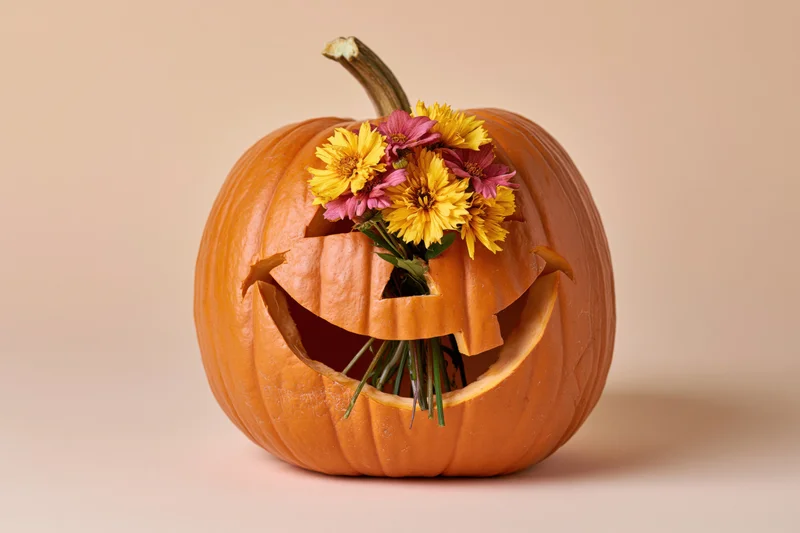 Carved pumpkin with a smiling face holding a bouquet of yellow and pink flowers against a beige background