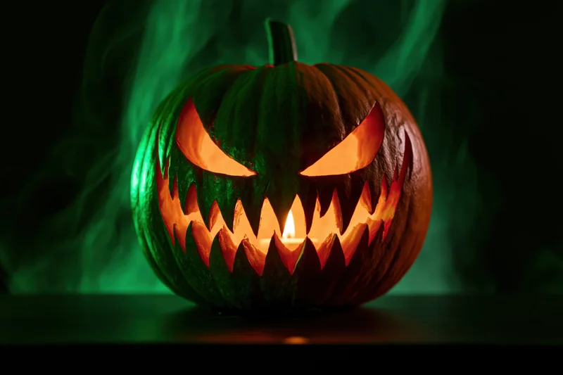 Carved pumpkin with jagged face lit by candle on dark surface with green smoke