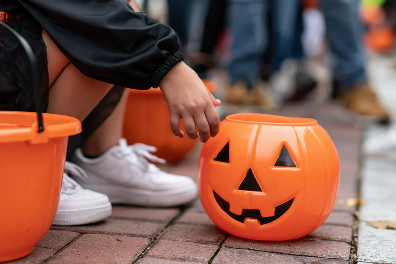 Child crouching on brick sidewalk reaching toward orange plastic pumpkin bucket during halloween trick or treat event