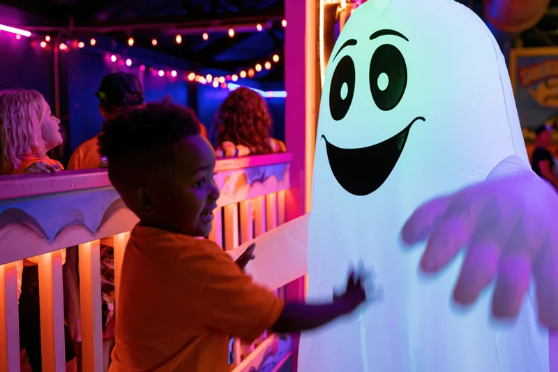 Child in orange shirt reaching toward glowing inflatable ghost at nighttime amusement park attraction