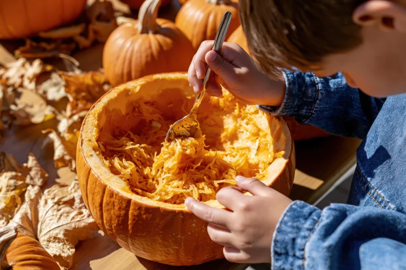 Child scooping out pumpkin flesh with a spoon at a table surrounded by autumn leaves and pumpkins