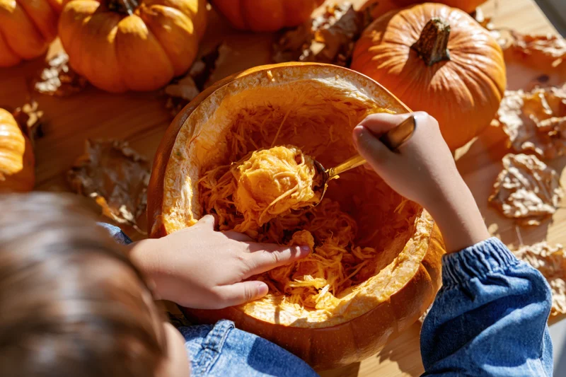 Child scooping out pumpkin flesh with a spoon on a wooden table surrounded by pumpkins