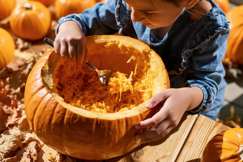 Child scooping out pumpkin flesh with a spoon on a wooden table surrounded by pumpkins