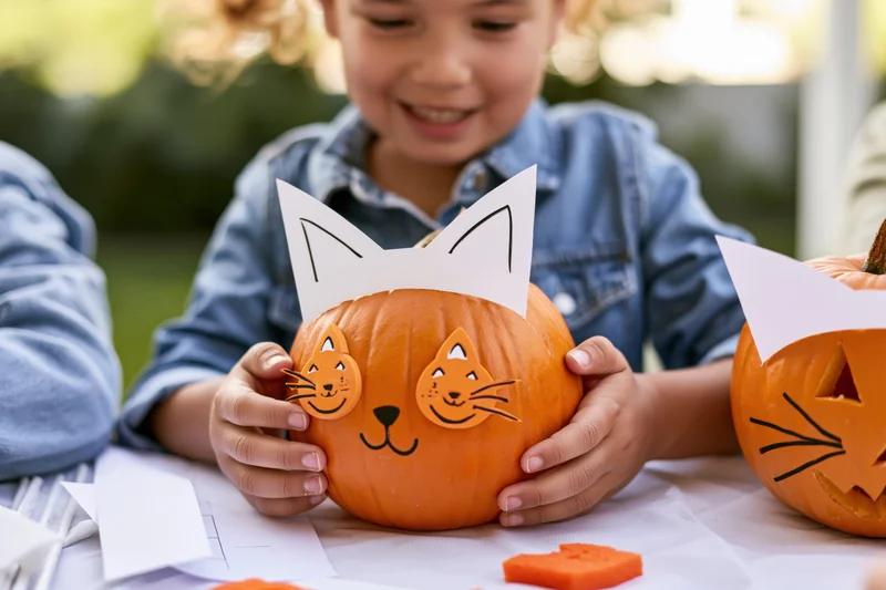 Child holding a pumpkin decorated with cat ears and face stickers at an outdoor table