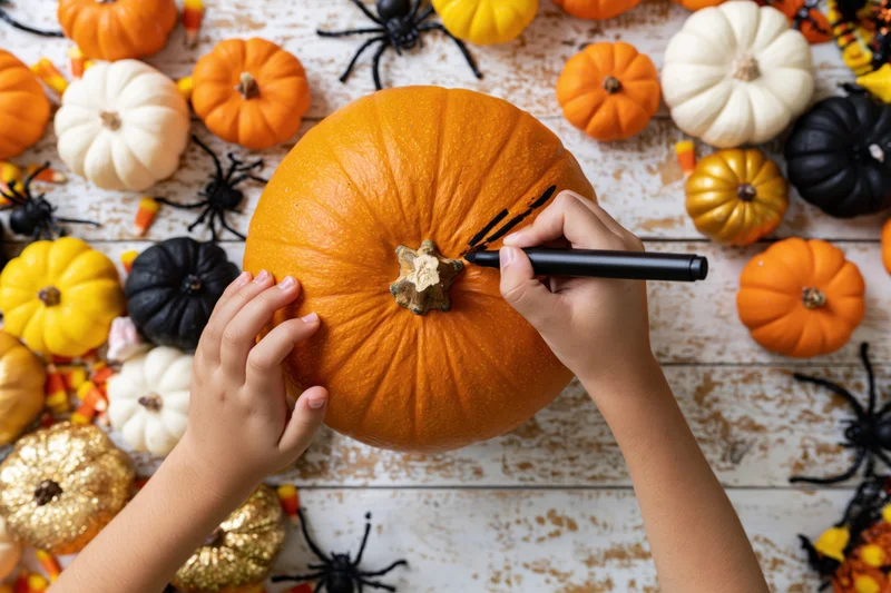 Child’s hands drawing on a pumpkin with a black marker on a decorated wooden table