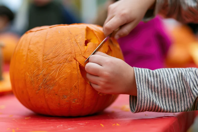 Child's hands carving a pumpkin on a red table with a small knife