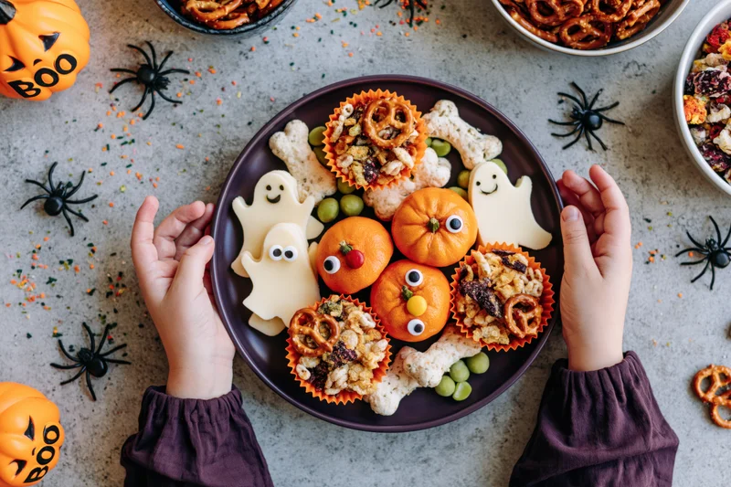 Child's hands holding a purple plate with halloween-themed snacks and decorations on a table