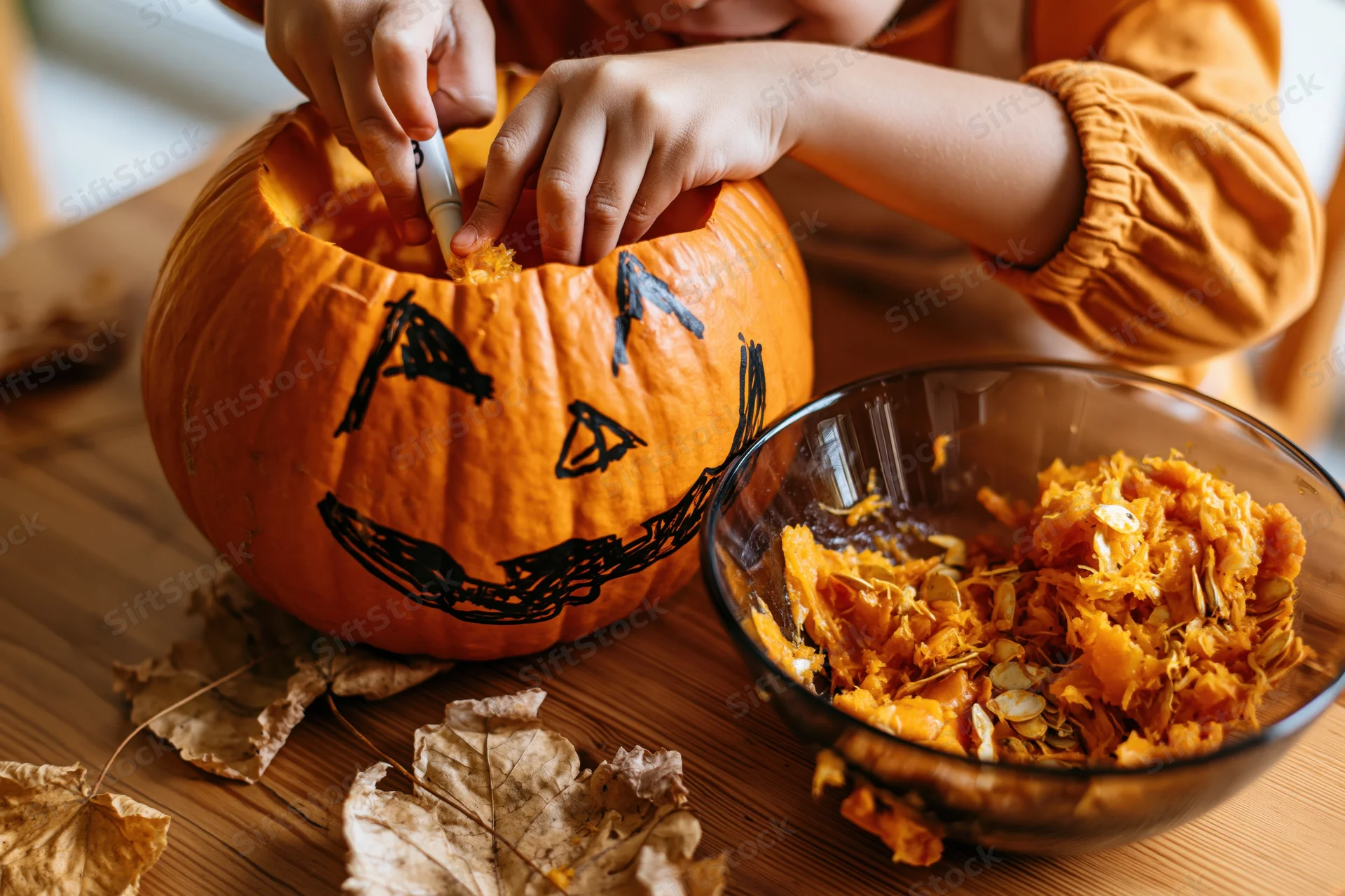 Child scooping out a carved pumpkin on a wooden table with dried leaves and a bowl of seeds nearby