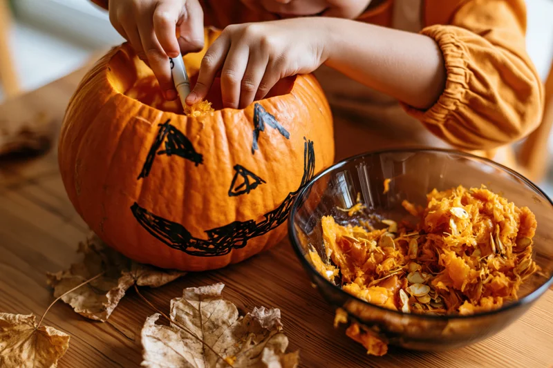 Child scooping out a carved pumpkin on a wooden table with dried leaves and a bowl of seeds nearby