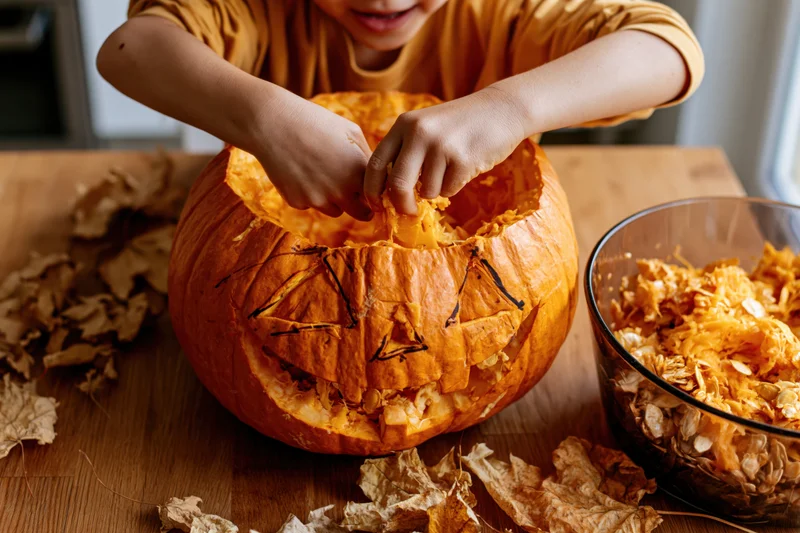 Child scooping out a carved pumpkin on a wooden table with dry leaves and a glass bowl of pulp nearby