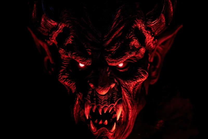Close-up of a demonic latex mask with glowing red eyes and sharp fangs in dark lighting