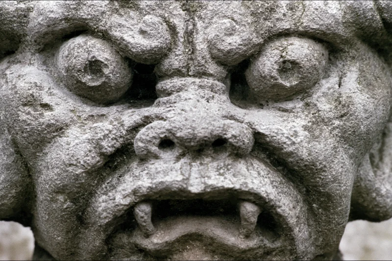 Close-up of a weathered stone gargoyle face with bulging eyes and fangs on a gothic building facade