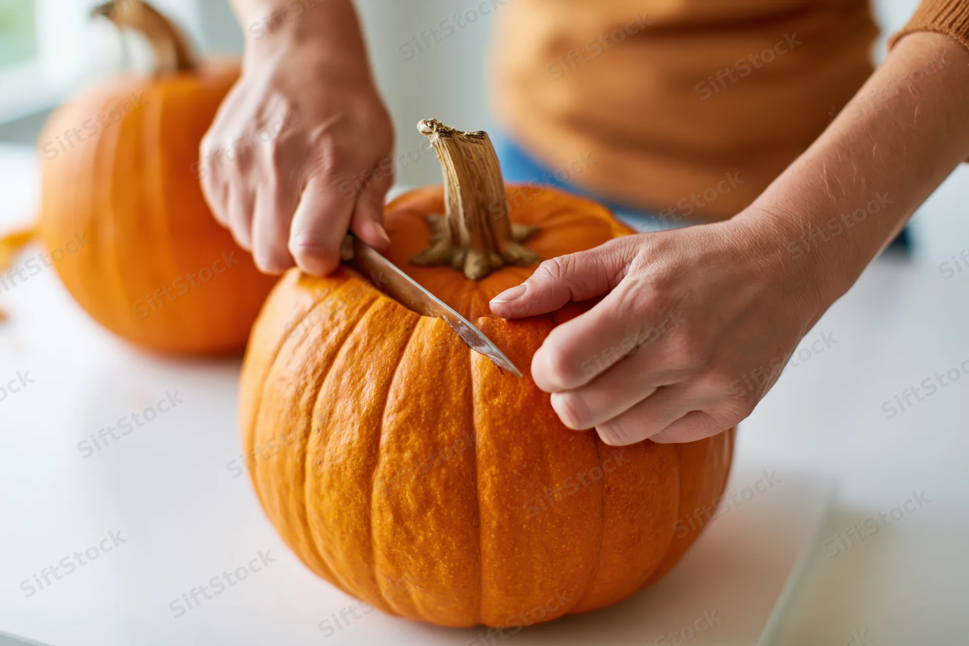 Hands carving a pumpkin on a white table with a small knife