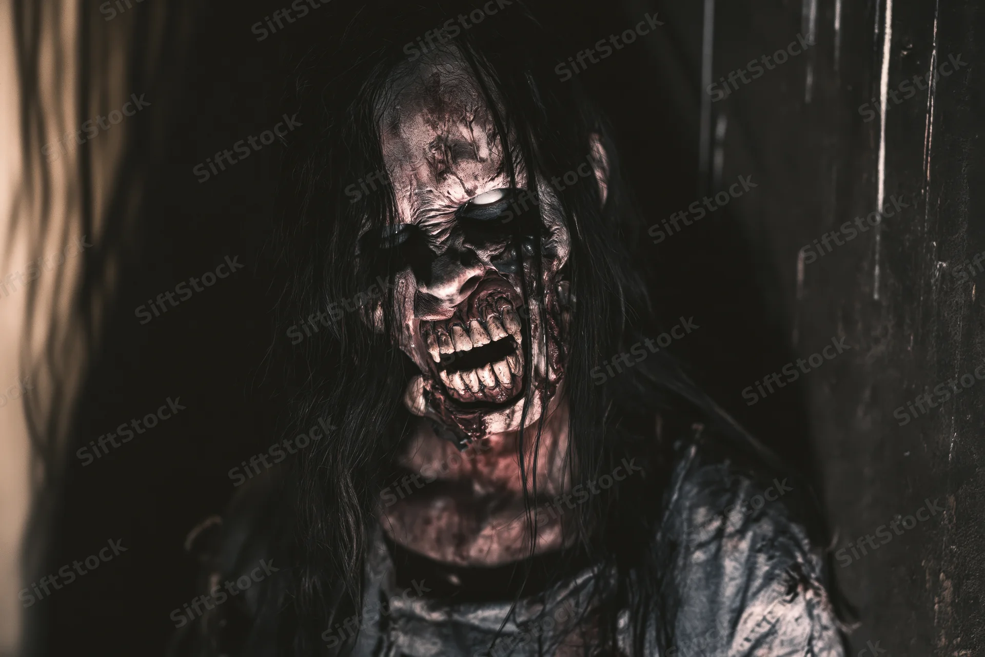 Close-up of a woman performer in elaborate zombie special effects makeup with decaying skin and bloodied teeth, standing in a dark, grimy setting