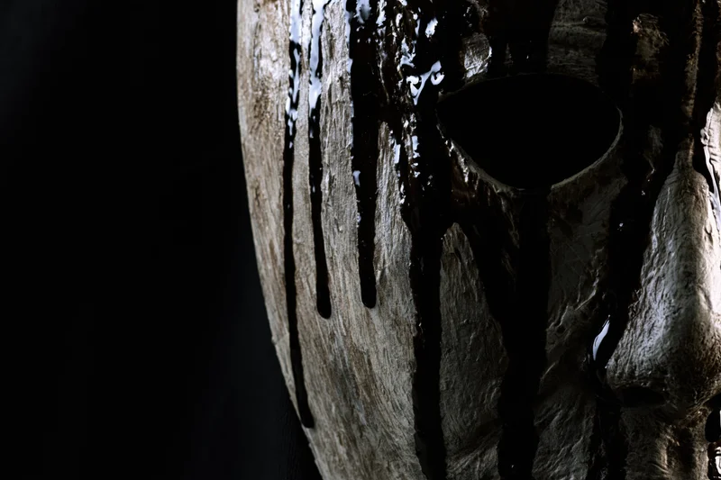 Close-up side profile of a weathered horror mask with black liquid dripping down its surface against a dark background