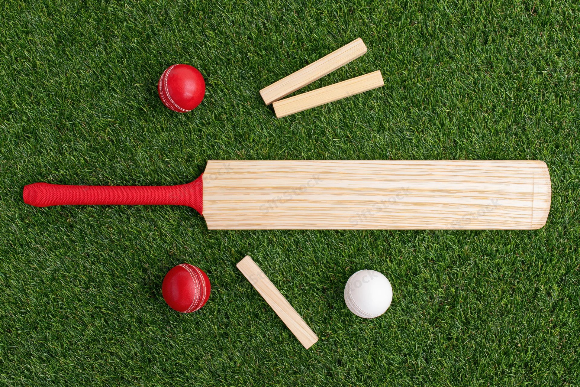 Cricket bat with red grip and balls and wickets arranged on artificial grass field