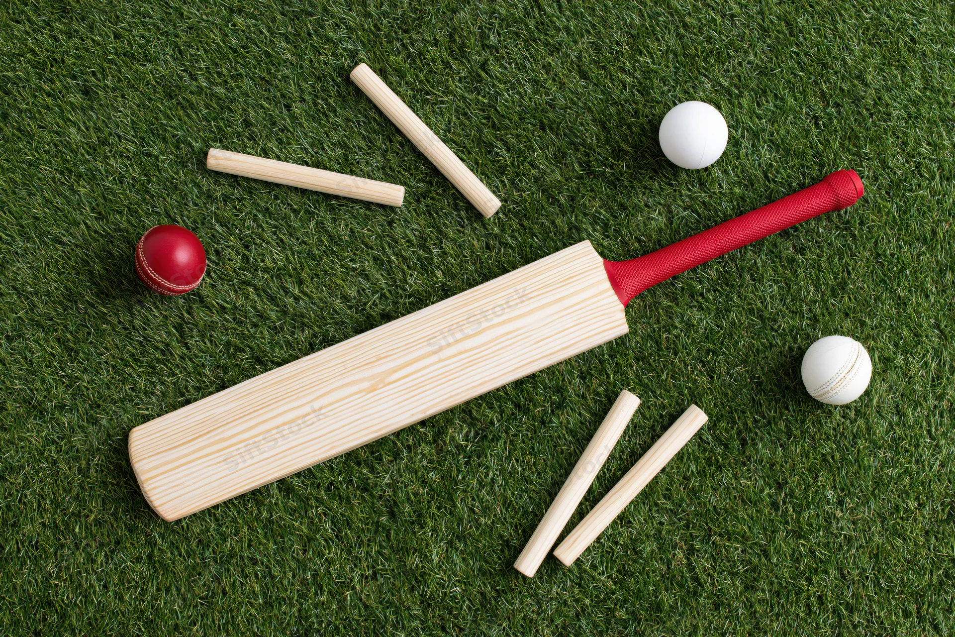 Cricket bat with red grip and balls lying on artificial grass field with wickets arranged around