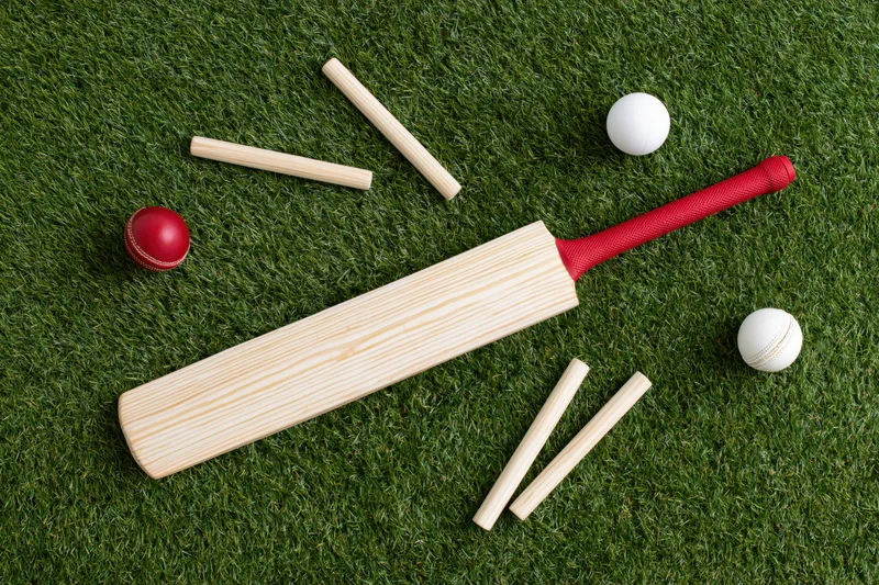 Cricket bat with red grip and balls lying on artificial grass field with wickets arranged around
