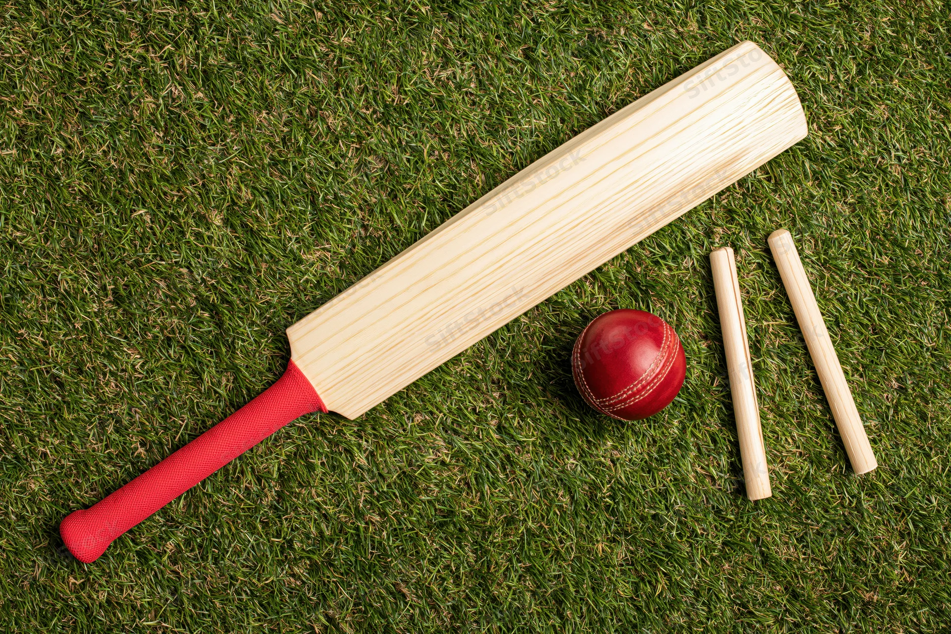 Cricket bat with red grip, red ball, and wooden wickets arranged on short green grass