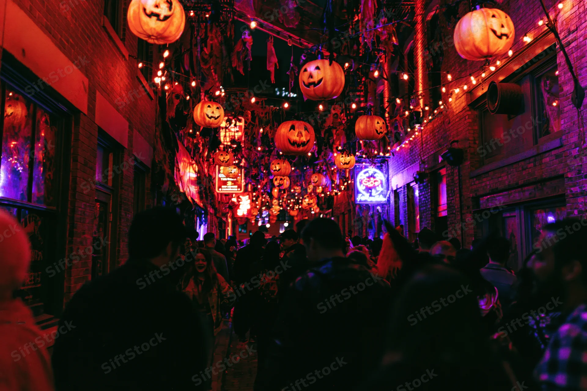 Crowd walking through narrow urban street decorated with glowing jack-o-lanterns and string lights at night