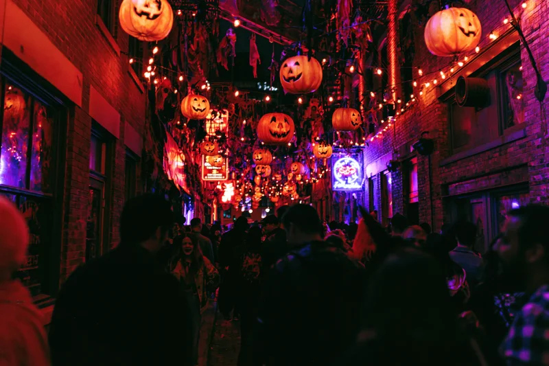 Crowd walking through narrow urban street decorated with glowing jack-o-lanterns and string lights at night