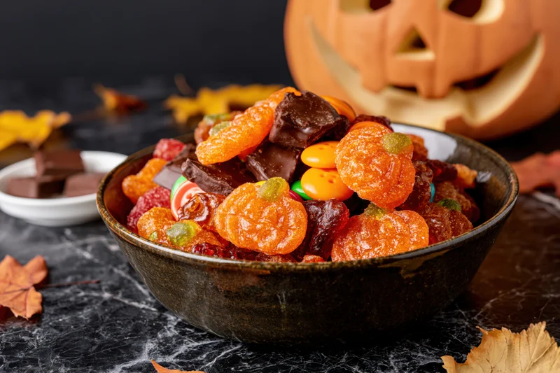 Dark ceramic bowl filled with halloween candy and chocolate pieces on a marble surface with carved pumpkin and autumn leaves nearby