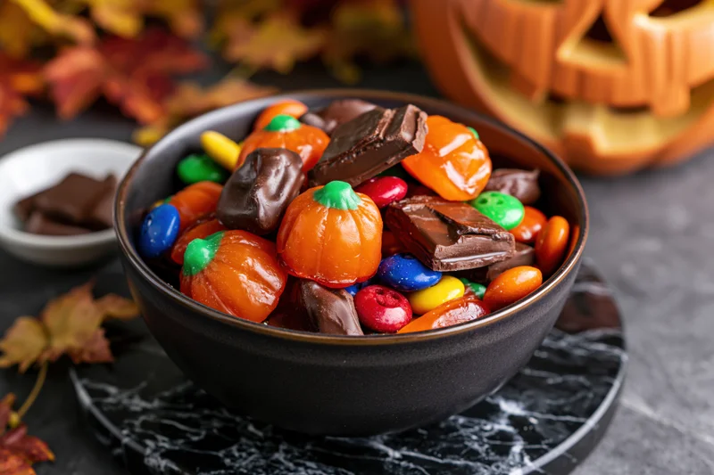 Dark ceramic bowl filled with halloween candy including pumpkin-shaped sweets and chocolate pieces on a marble plate with autumn leaves and a jack-o-lantern in the background