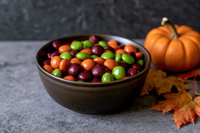 Bowl of orange green and deep red candy on gray surface with pumpkin and autumn leaves