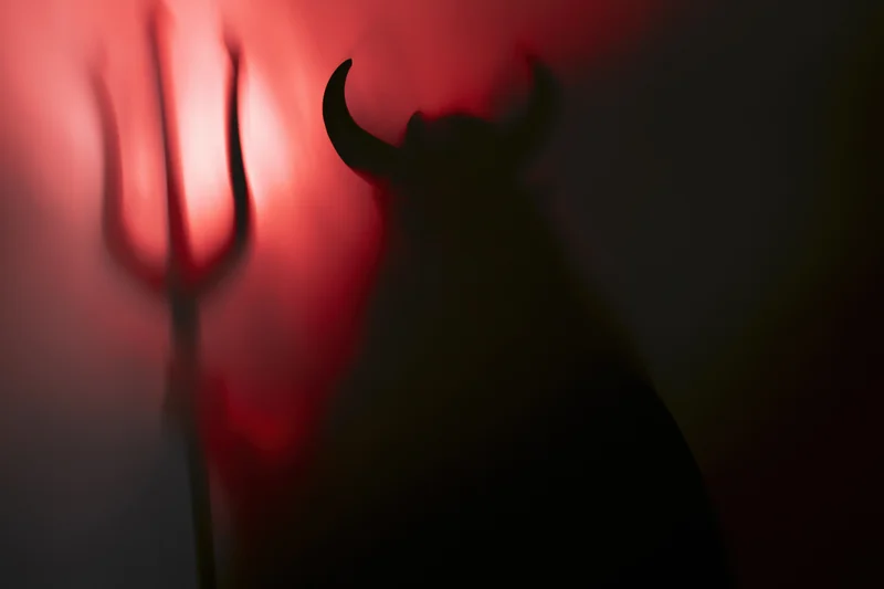 Dark silhouette of a devil figure with horns holding a trident against red-lit smoke