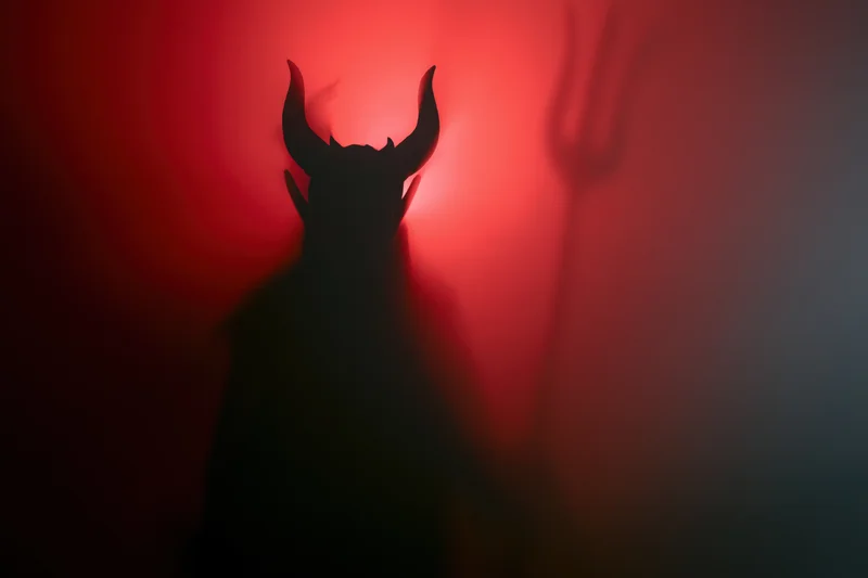 Dark silhouette of a devil figure with horns holding a trident against red glowing background