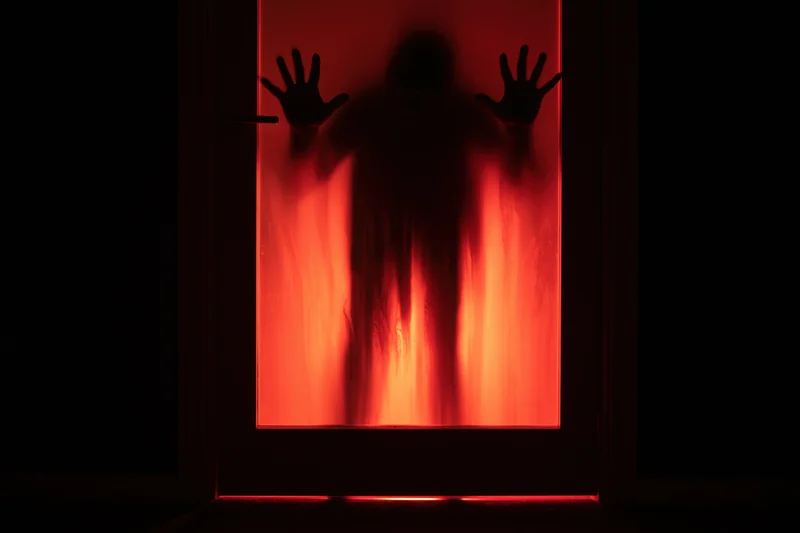 Dark silhouette of a person pressing hands against frosted glass door glowing red in the dark