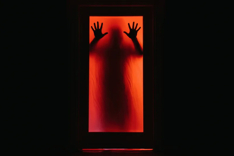 Dark silhouette of a figure pressing hands against a frosted door lit with red light