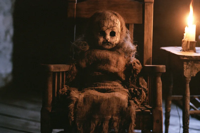 Decayed antique doll with cracked pale face and hollow eyes sitting in wooden chair beside lit candle in dim room