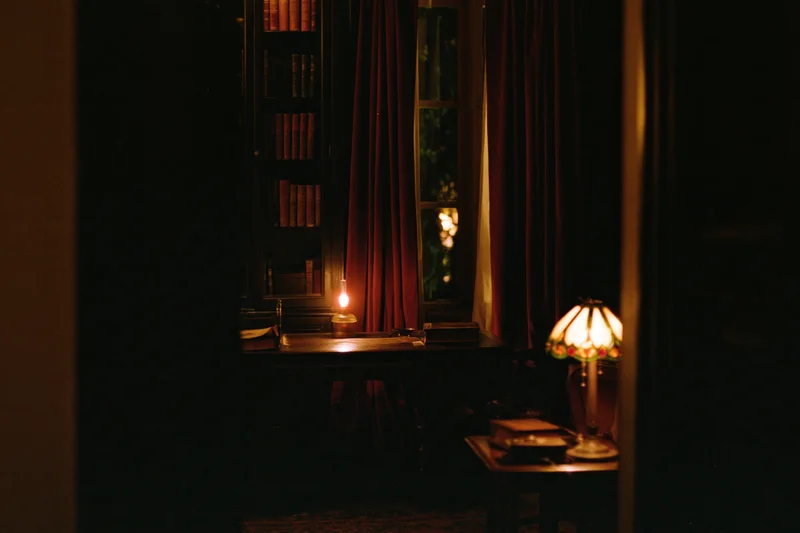 Dimly lit vintage study room with wooden desk, lamp, bookshelf and red curtains at night