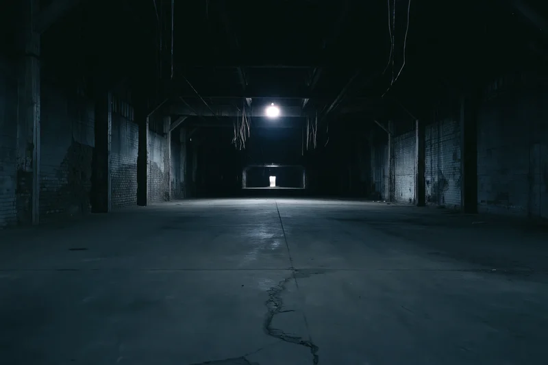 Empty abandoned industrial warehouse interior with single overhead light casting long shadows on cracked concrete floor